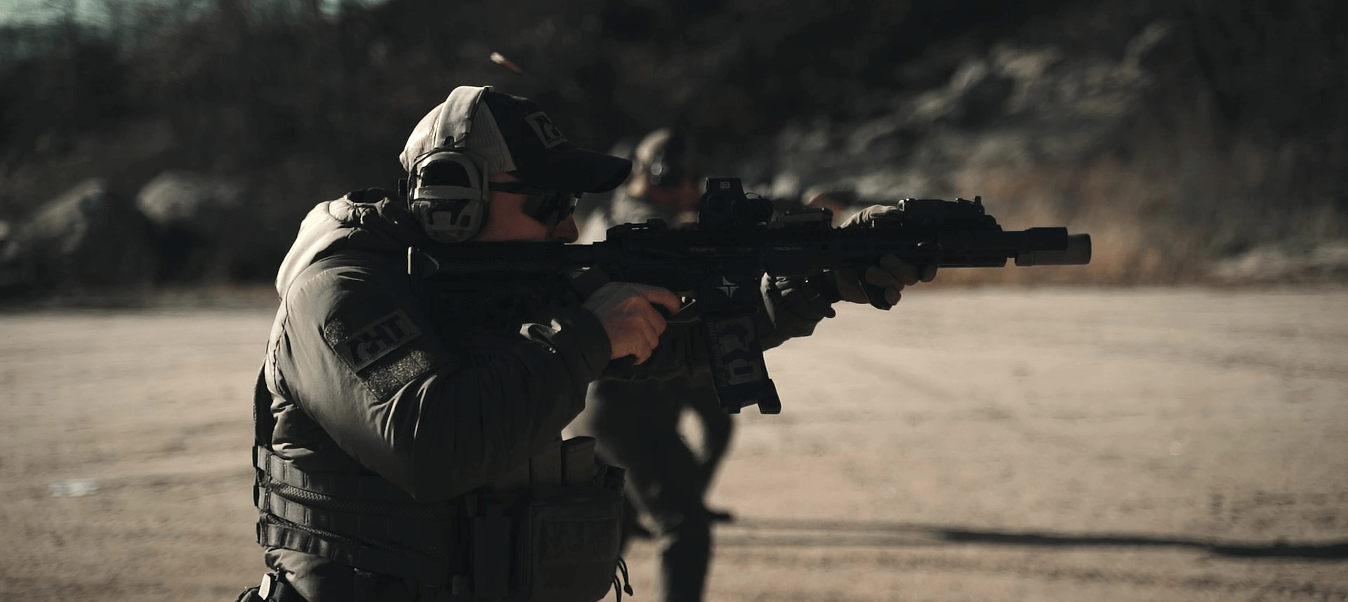 A person wearing tactical gear and headphones aims a rifle outdoors on a dirt surface with another similarly dressed individual visible in the background.